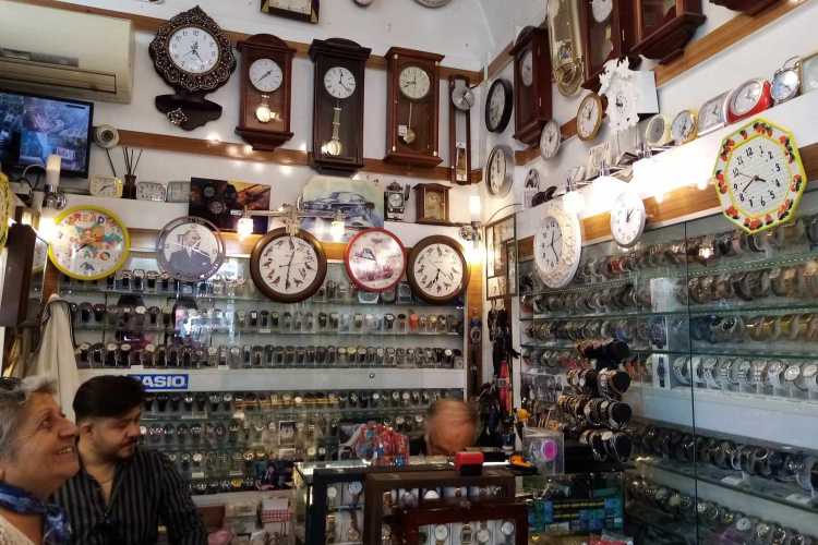 Watch repairman sitting behind a counter inside a watch shop, repairing a wristwatch, with many clocks displayed on the walls.