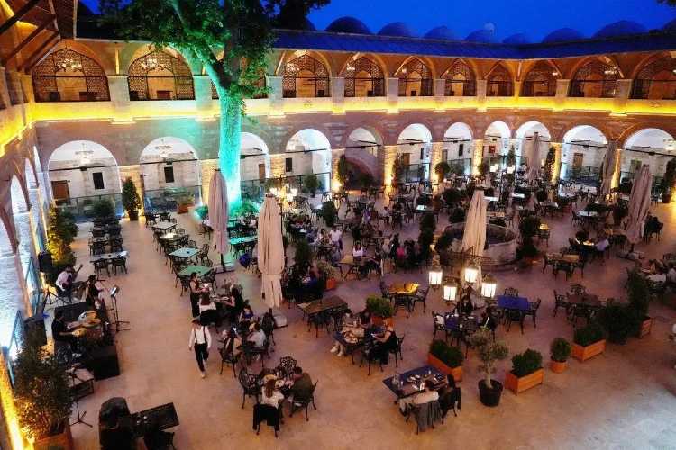 Inner courtyard of Rüstem Pasha Caravanserai in Edirne, showing a square open-air garden at evening time with warm romantic lighting, surrounded by two-story Ottoman rooms and arches, with people sitting at tables in the illuminated courtyard.