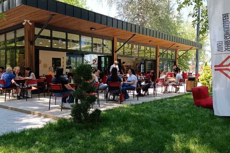 People sitting at outdoor café tables in Edirne Millet Bahçesi, a green riverside park with a relaxed social atmosphere
